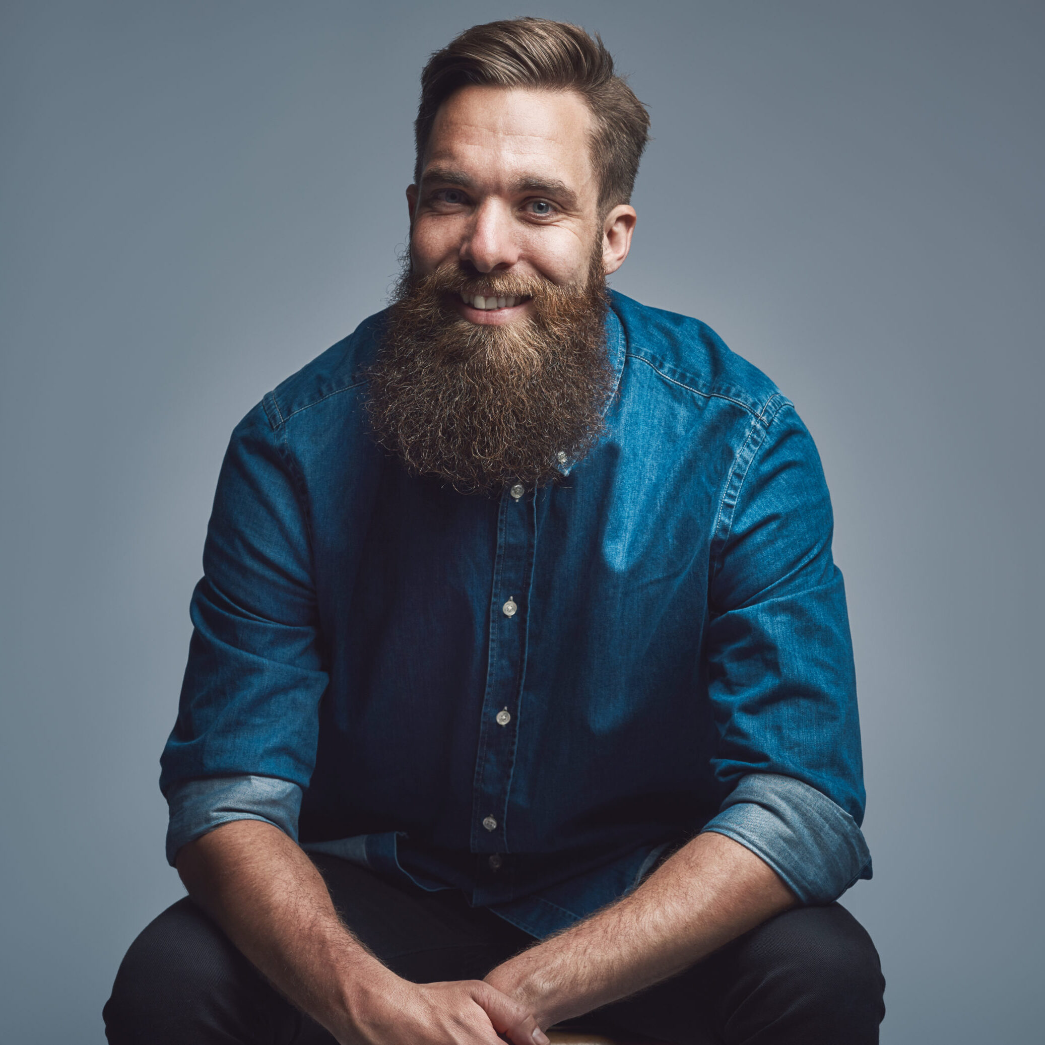 Single sitting young handsome smiling man in blue denim shirt and beard over gray background