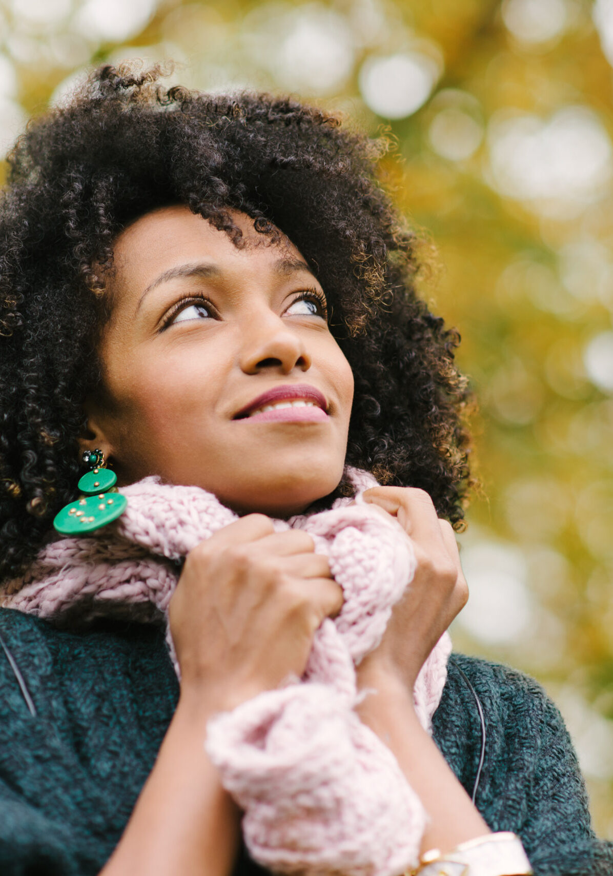 Black pensive woman wearing warm scarf looking up to the autumn trees at the park.