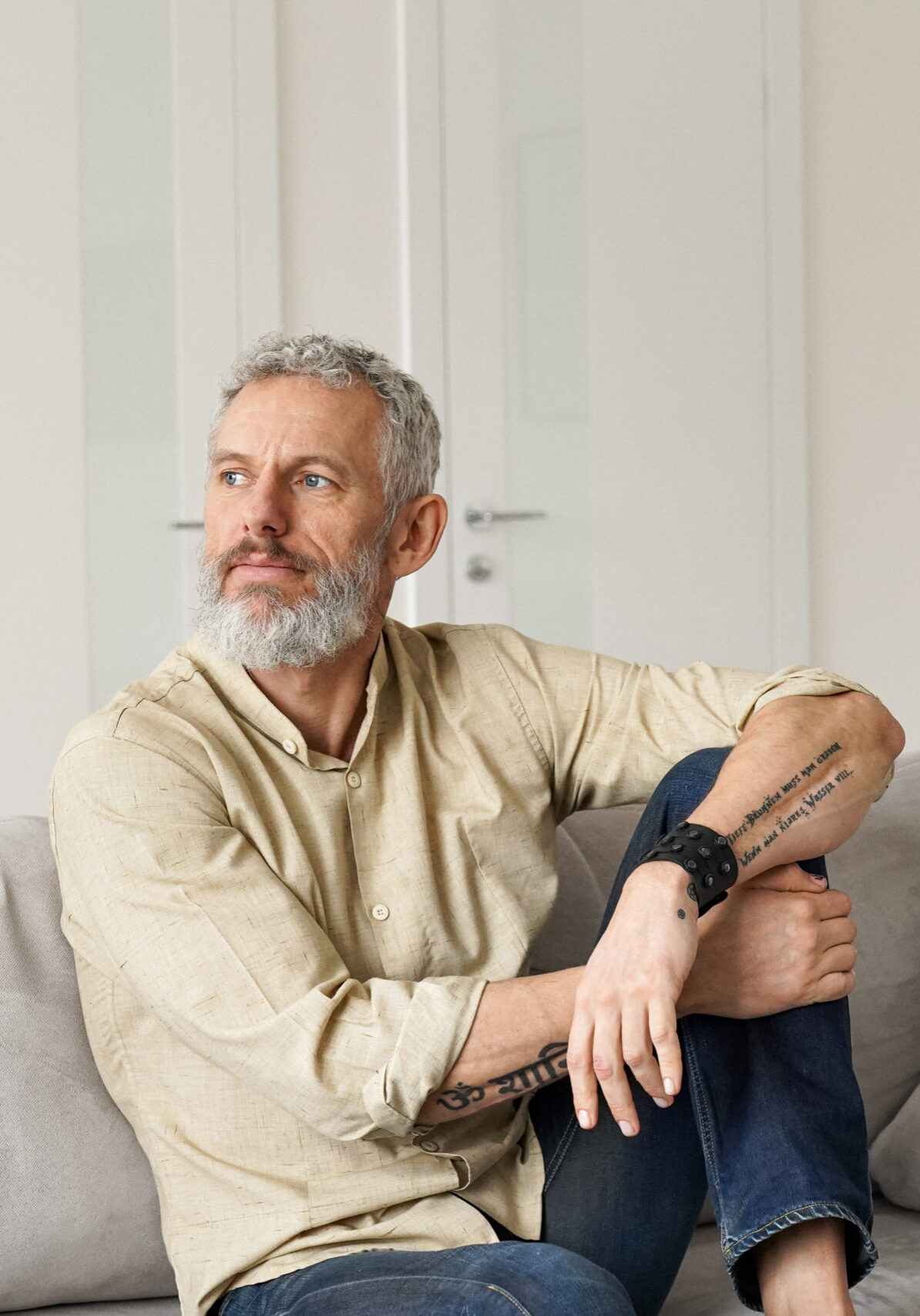 Relaxed mature older thoughtful tattooed hipster man relaxing sitting on sofa and thinking at home. Pensive calm stylish mid aged single bearded guy resting on couch at home looking away, reflecting.