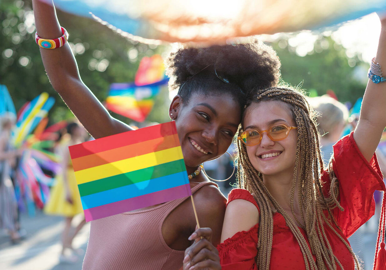 A young black female leans into a white female as they hold a Pride flag in celebration of Pride Month.